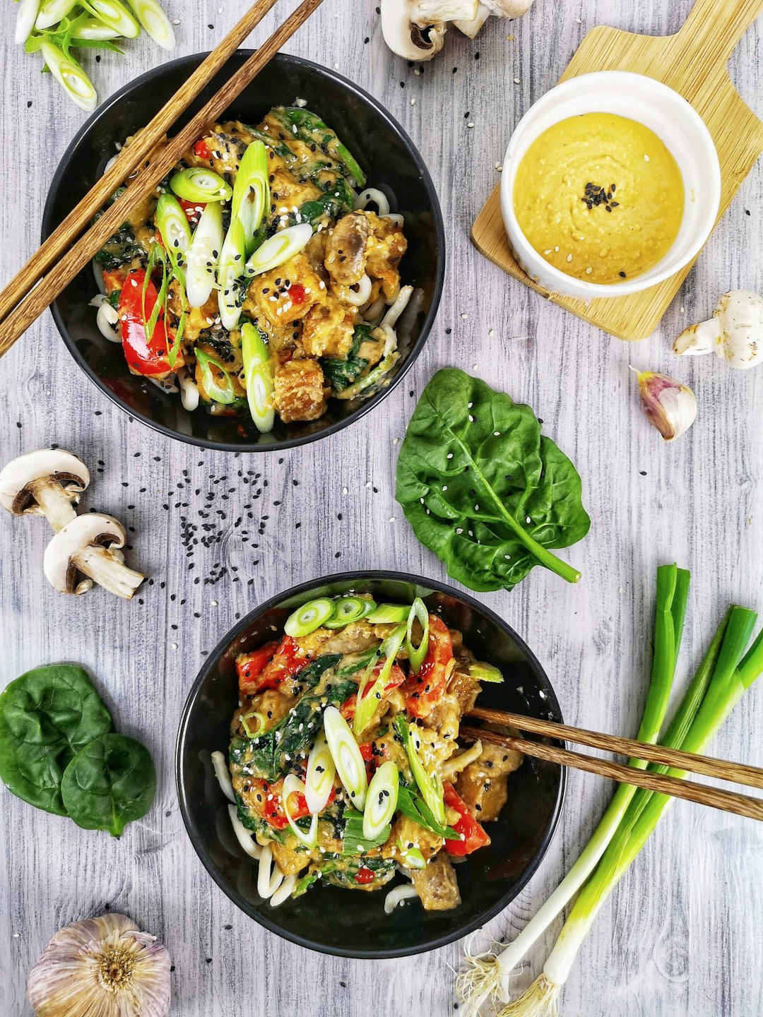 Two bowls of noodles with chicken, peppers, and spinach on wood.